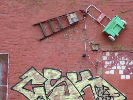 Kunst an der Wand Das Foto zeigt eine rote, verfallene Wand. Teilweise ist die Farbe abgeblättert. Unten ist ein Graffity mit gelben, schwarz umrandeten Zeichen zu sehen. Darüber ist an der Wand eine schwarze Leiter mit vier Stufen befestigt. Rechts neben der Leiter ist eine Sackkarre befestigt mit weißen Rädern und einem grünen Kunststoffkorb.