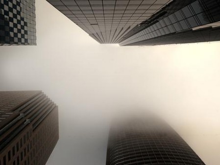 Das Foto zeigt vier Wolkenkratzer, die nebeneinander stehen und von unten fotografiert sind. Die Spitze des einen Wolkenkratzers geht im Nebel unter, die Spitzen der anderen kann man ahnen. Farben sind kaum wahrzunehmen, weil alles im Grau des Nebels liegt, durch den kaum Licht dringt.