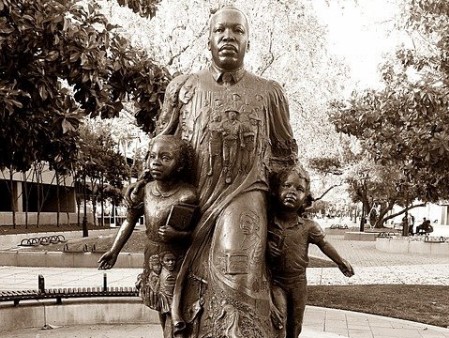 Martin Luther King Das Schwarzweiß-Foto zeigt eine Statue von Martin Luther King. Er schreitet nach vorn aus, das linke Bein vorgestellt. Sein Gesicht ist ernst und entschlossen. An beiden Armen führt er zwei Kinder mit sich, am rechten Arm ein Mädchen und am linken Arm einen etwas kleineren Jungen. Auf seiner Brust sind Männer und Frauen eingraviert, die ebenfalls nach vorn ausschreiten und offenbar mit ihm unterwegs sind. Daneben sieht man weitere Gesichter. Auf seinem linken Bein sind weitere eingravierte Menschen zu sehen. Die Statue steht auf einem Podest mit der Aufschrift "I have a dream". Im Hintergrund sind die Bäume eines Parks und einige Bänke zu sehen.