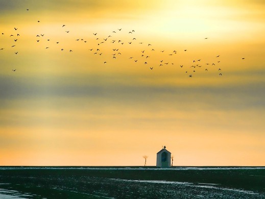 Das Foto zeigt eine ebene Landschaft mit einer kleinen Kapelle in der Ferne. Der Abendhimmel ist orangefarben, und ein Vogelschwarm ist zu sehen. Alles strahlt Freude und Glück aus - so, wie der christliche Glaube tiefe Freude und seelisches Glück bedeutet.